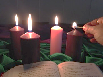 4 Advent candles on green cloth with a Bible in the foreground. From left to rights: 2 lit purple candles, 1 lit pink candle and a person lighting the far right purple candle with a match.
